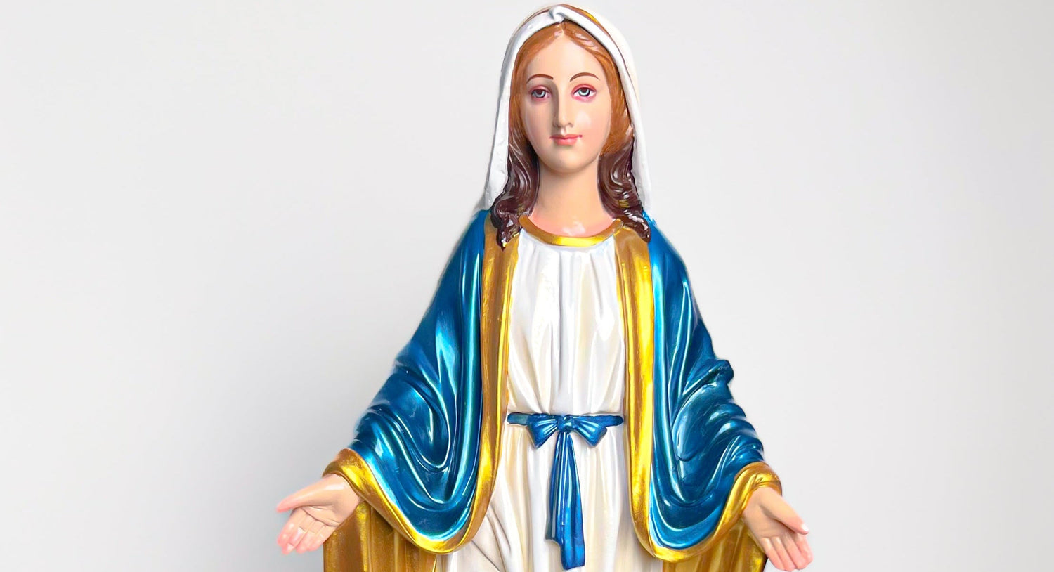 A religious figurine of the Virgin Mary with outstretched hands, wearing a white dress, a blue mantle with gold trim, and a white head covering, set against a plain light gray background.